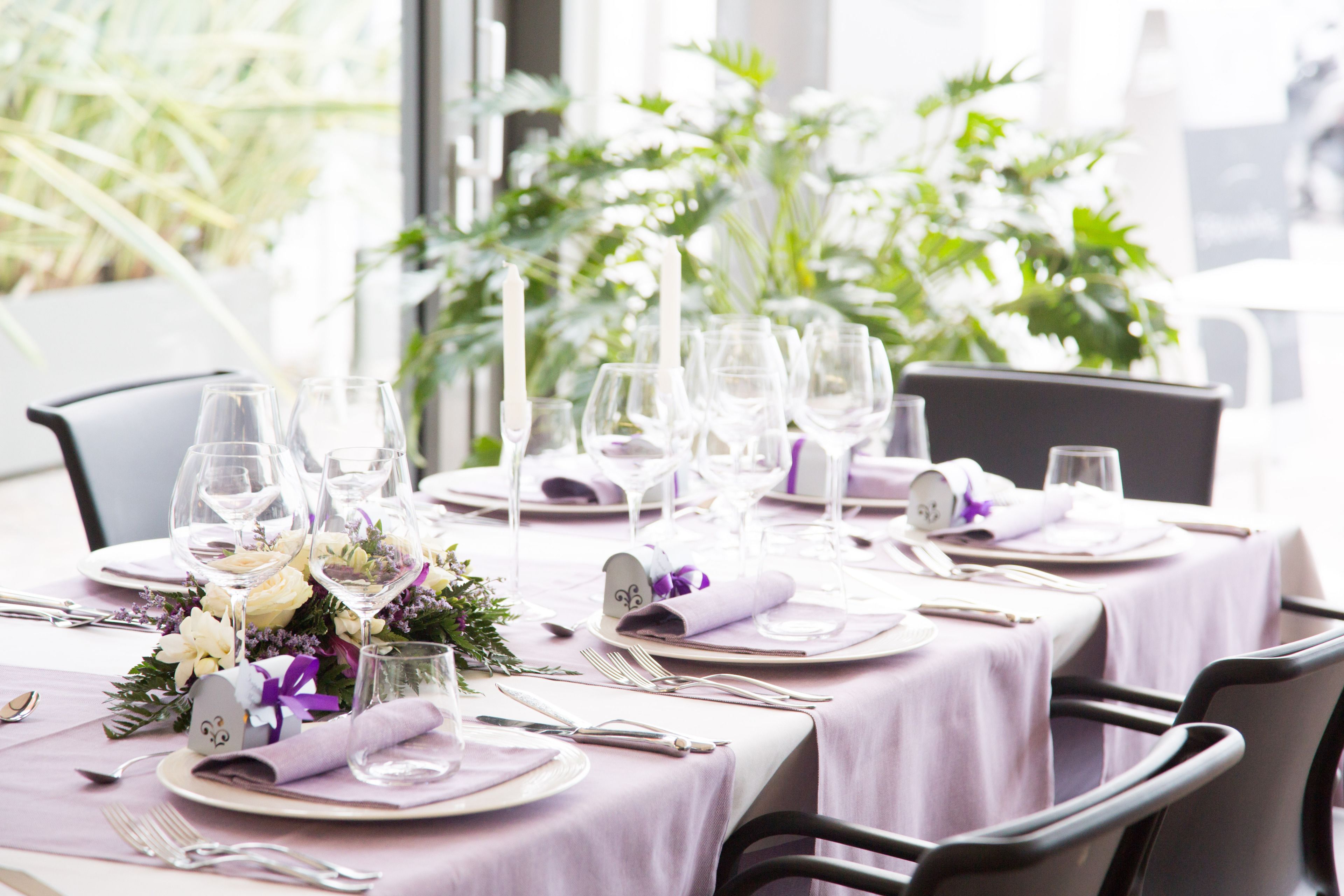 Una mesa dispuesta para un evento gastronómico formal, con manteles blancos y púrpuras, cristalería transparente y un centro de mesa floral.