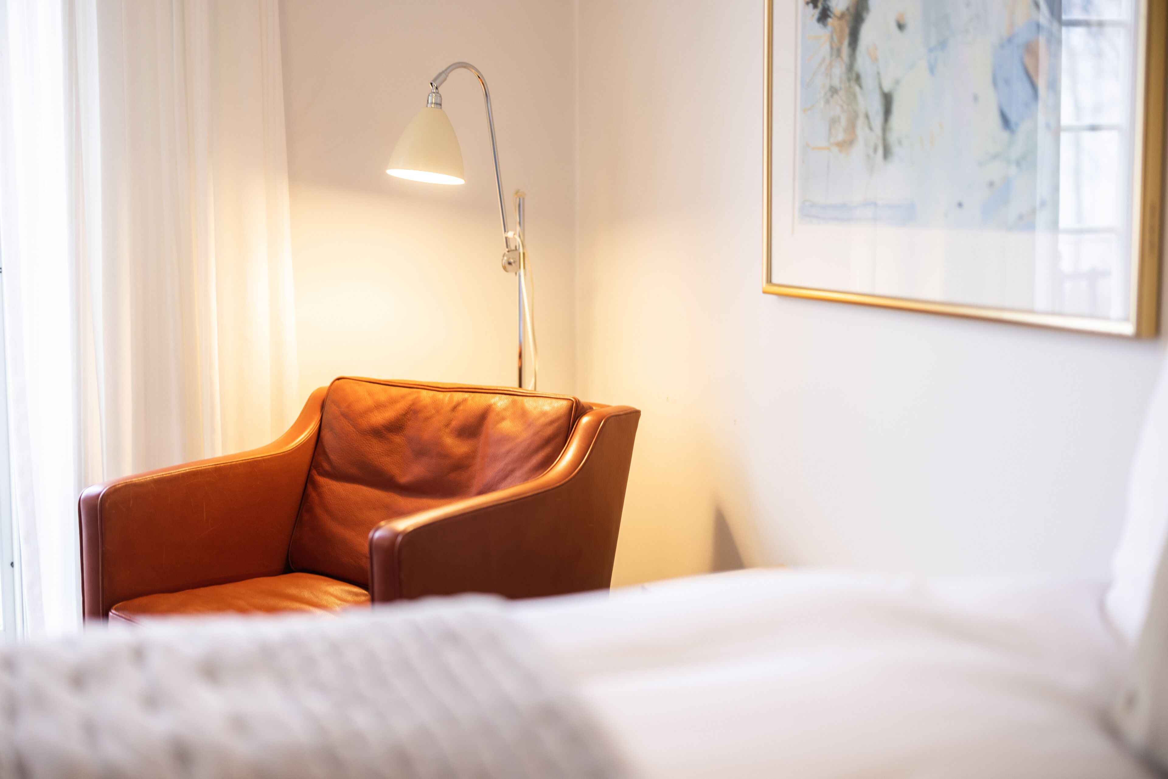 Habitación de hotel con un sillón de cuero marrón, una cama blanca, una lámpara blanca y un cuadro abstracto enmarcado en la pared.