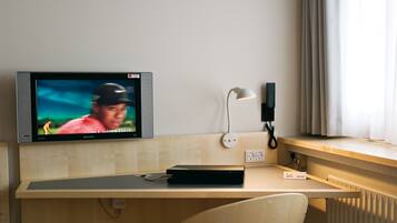 A hotel room with a wooden desk, a flat-screen TV, a desk lamp, a chair, and a window with curtains.