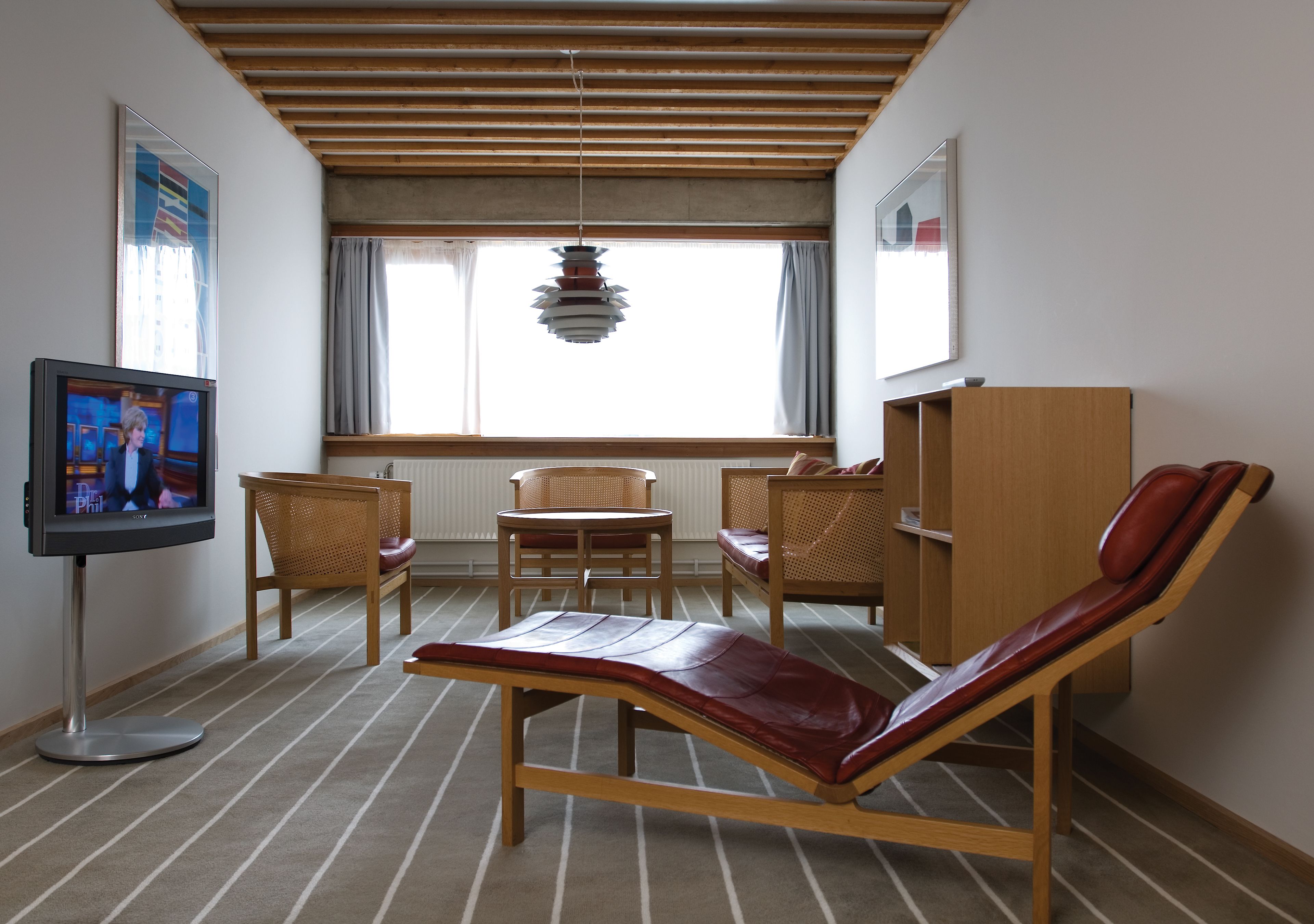 Una habitación con televisor, una butaca reclinable, dos sillas, un armario de madera y una ventana con cortinas.