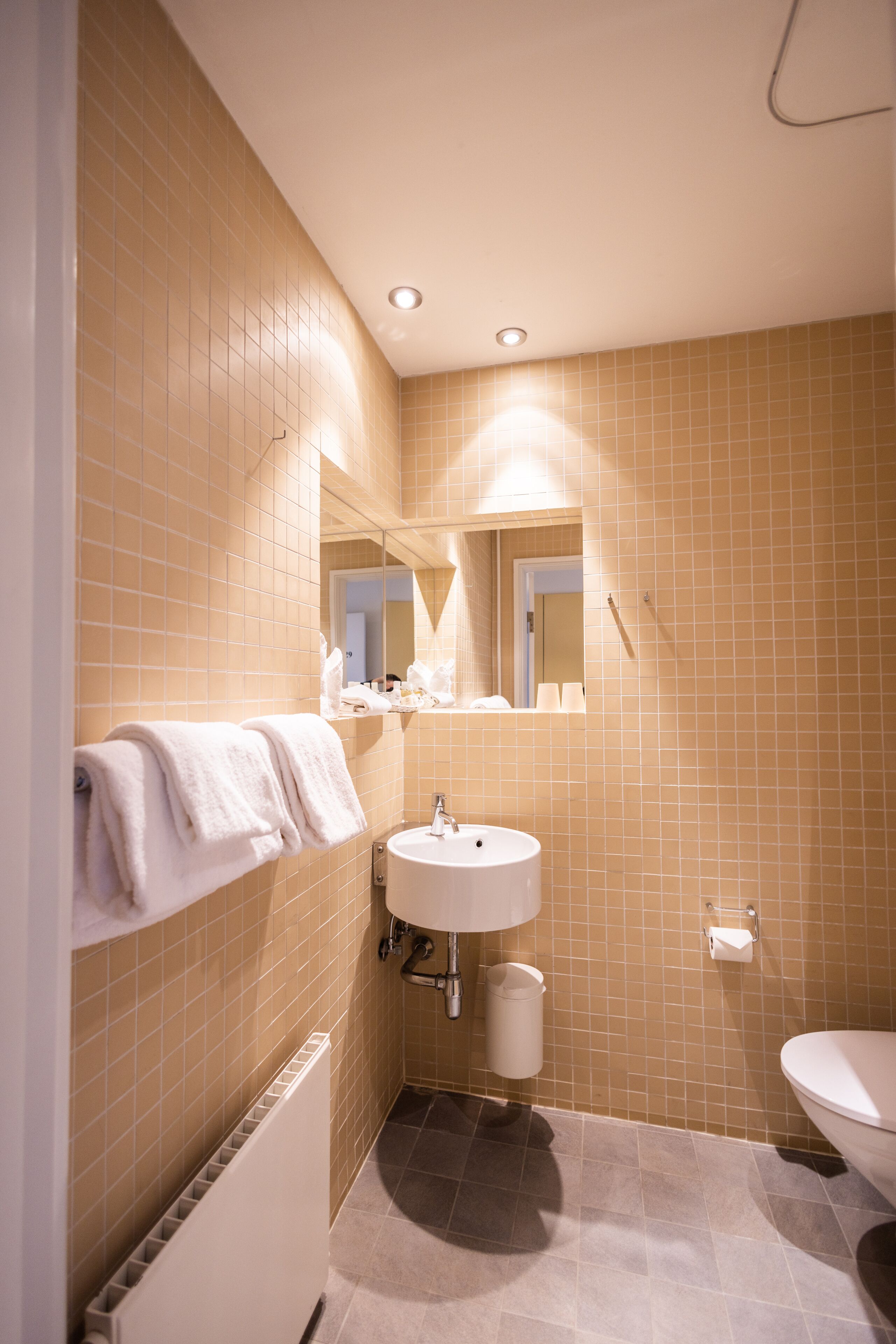 Een badkamer met behang van beige tegels, een witte wastafel, een spiegel boven de wastafel, een handdoekhouder en een toilet.