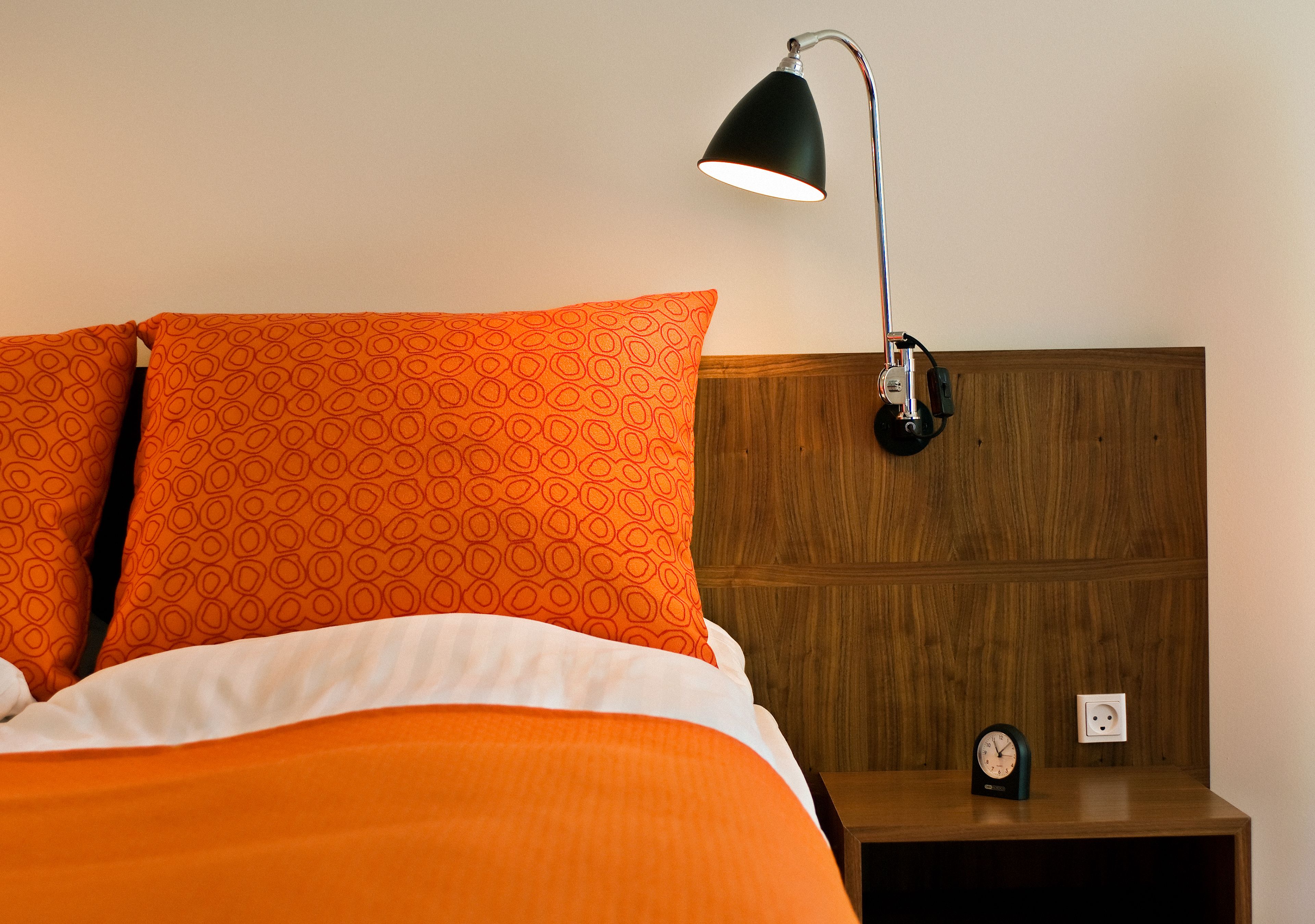 A bed with orange bedding, a black desk lamp, and a small clock on a wooden nightstand.