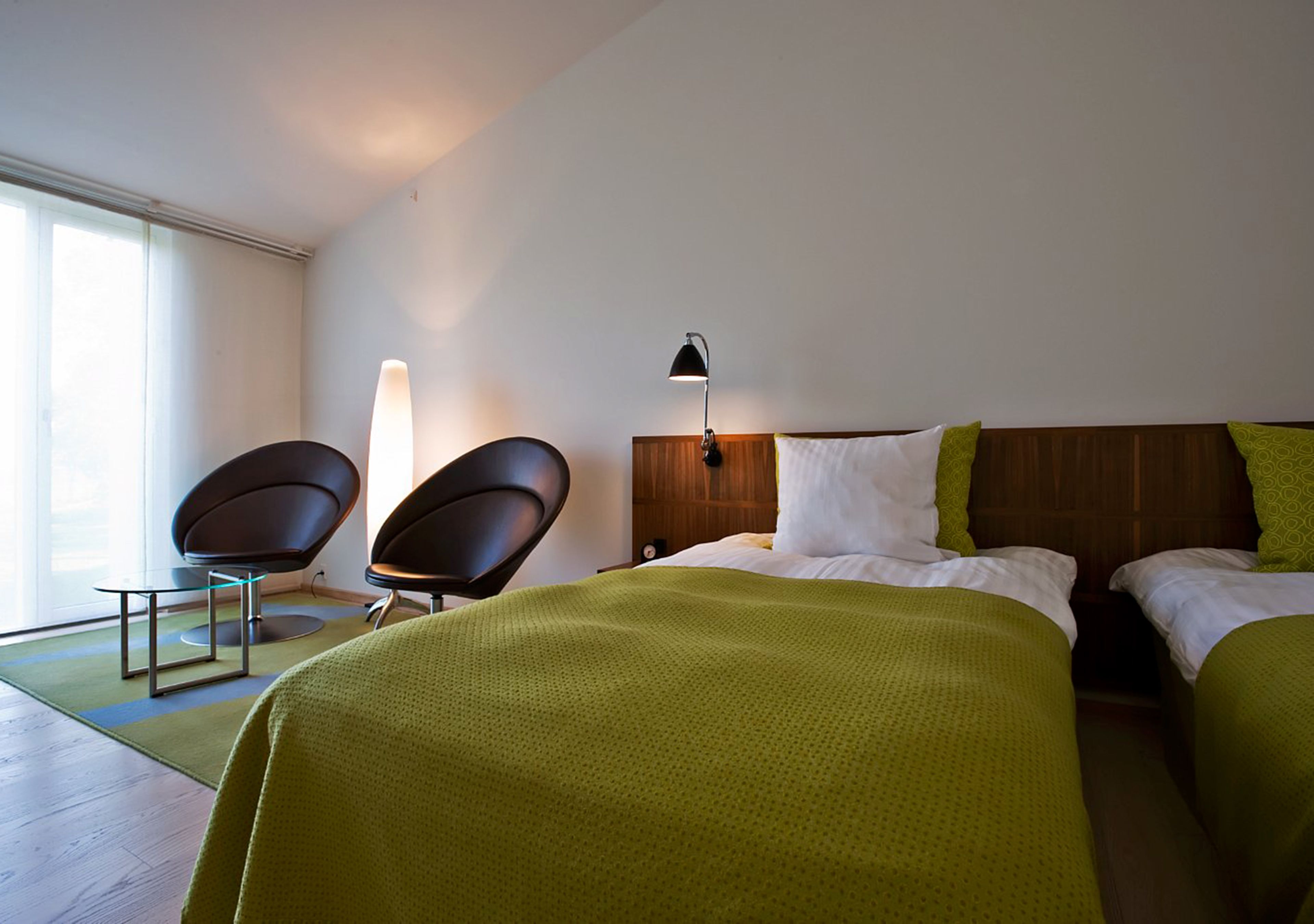 A hotel room with two beds, a desk, two chairs, a nightstand, and a lamp.