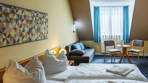 Une chambre d’hôtel comprenant un lit, un canapé, une petite table avec des chaises, une œuvre d’art murale et une fenêtre avec des rideaux.