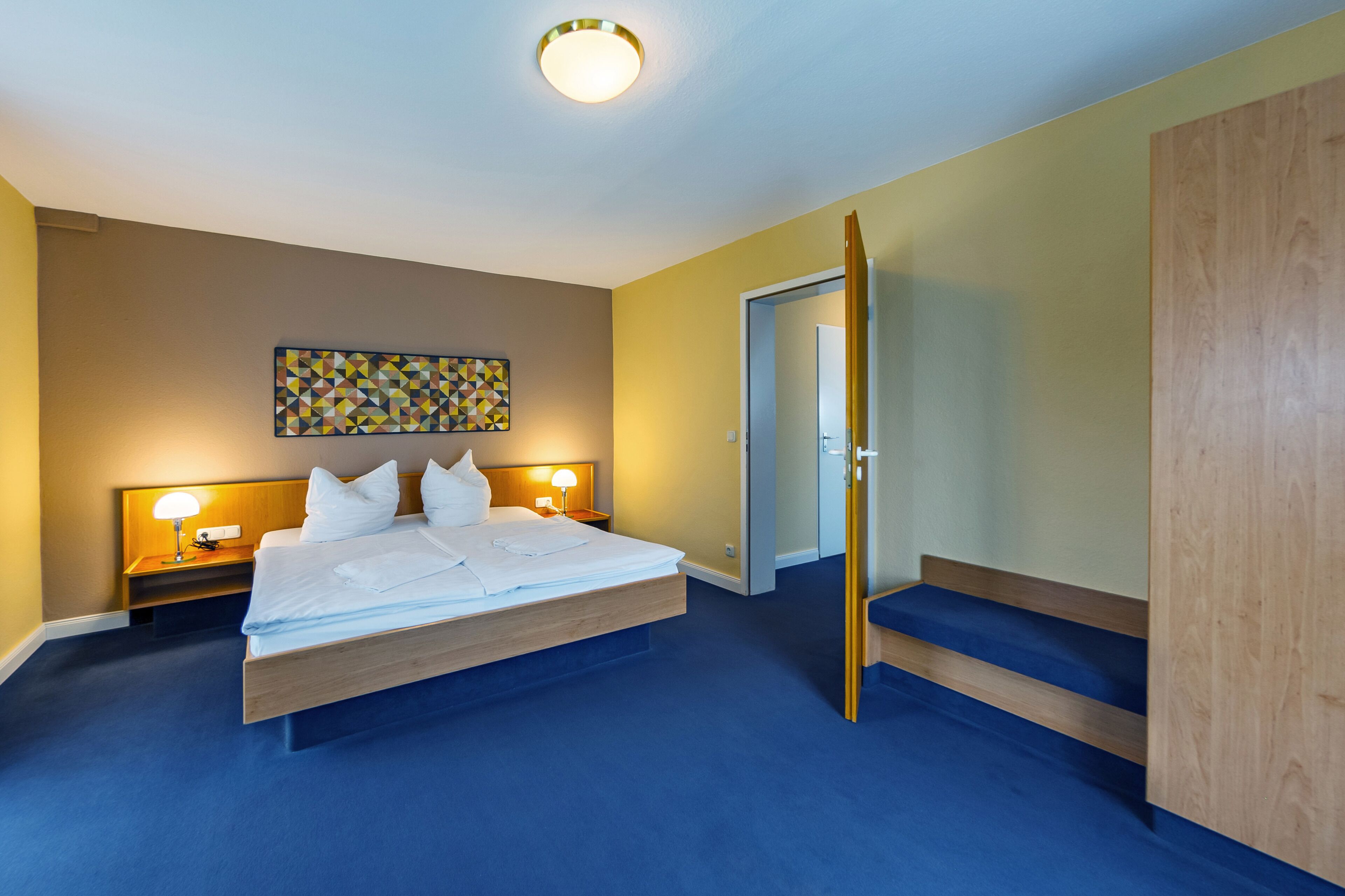 Une chambre d’hôtel avec un grand lit, deux tables de chevet avec des lampes, une armoire en bois et un tableau abstrait aux couleurs vives accroché au mur.