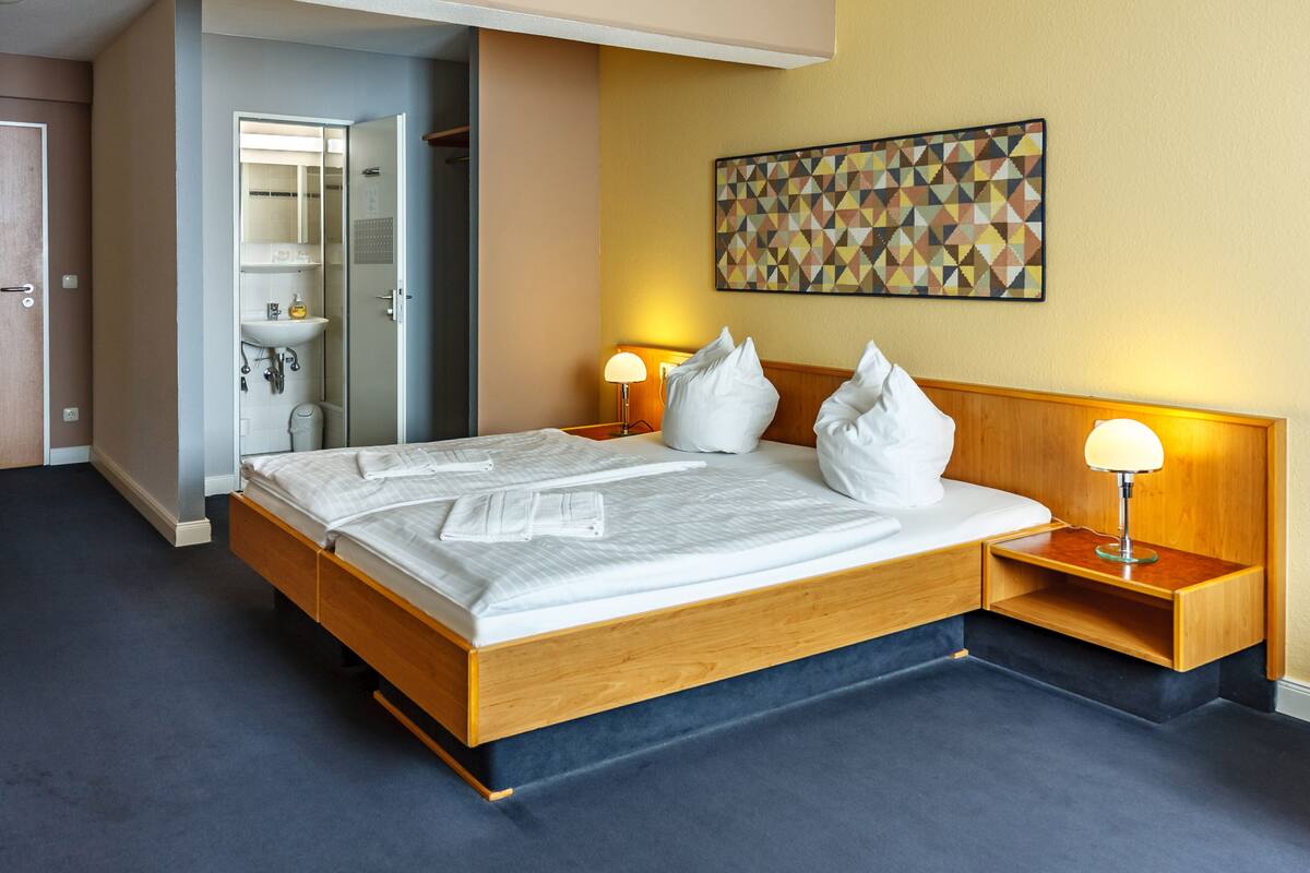 A hotel room with a wooden bed, bedside lamps, a wall-mounted geometric artwork, and a bathroom visible through an open door.