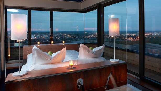 A modern bedroom with a large bed, bedside lamps, and a view of a cityscape through large windows. - The Seven Hotel (Esch-sur-Alzette)