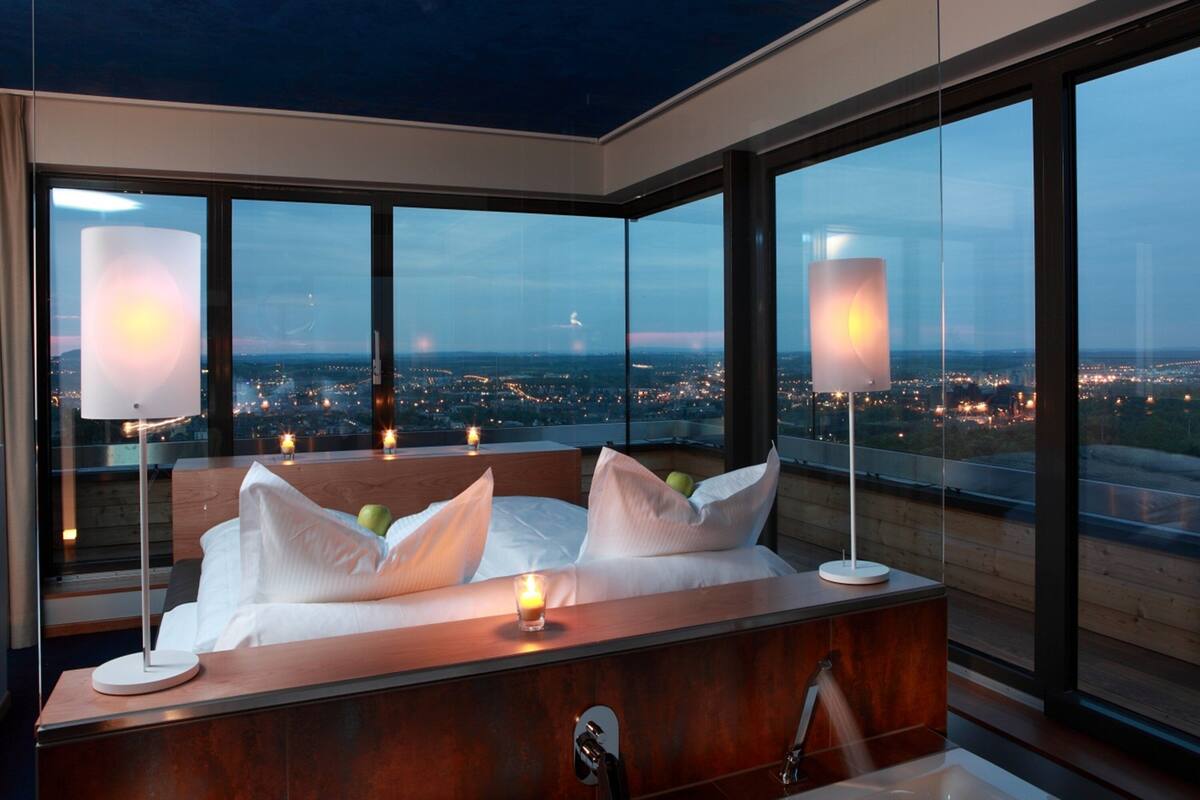 A modern bedroom with a large bed, bedside lamps, and a view of a cityscape through large windows.