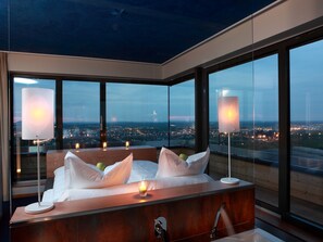 A modern bedroom with a large bed, bedside lamps, and a view of a cityscape through large windows. - The Seven Hotel (Esch-sur-Alzette)