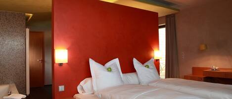 Habitación de hotel con una cama grande, dos mesitas de noche, una lámpara y vista al pasillo.
