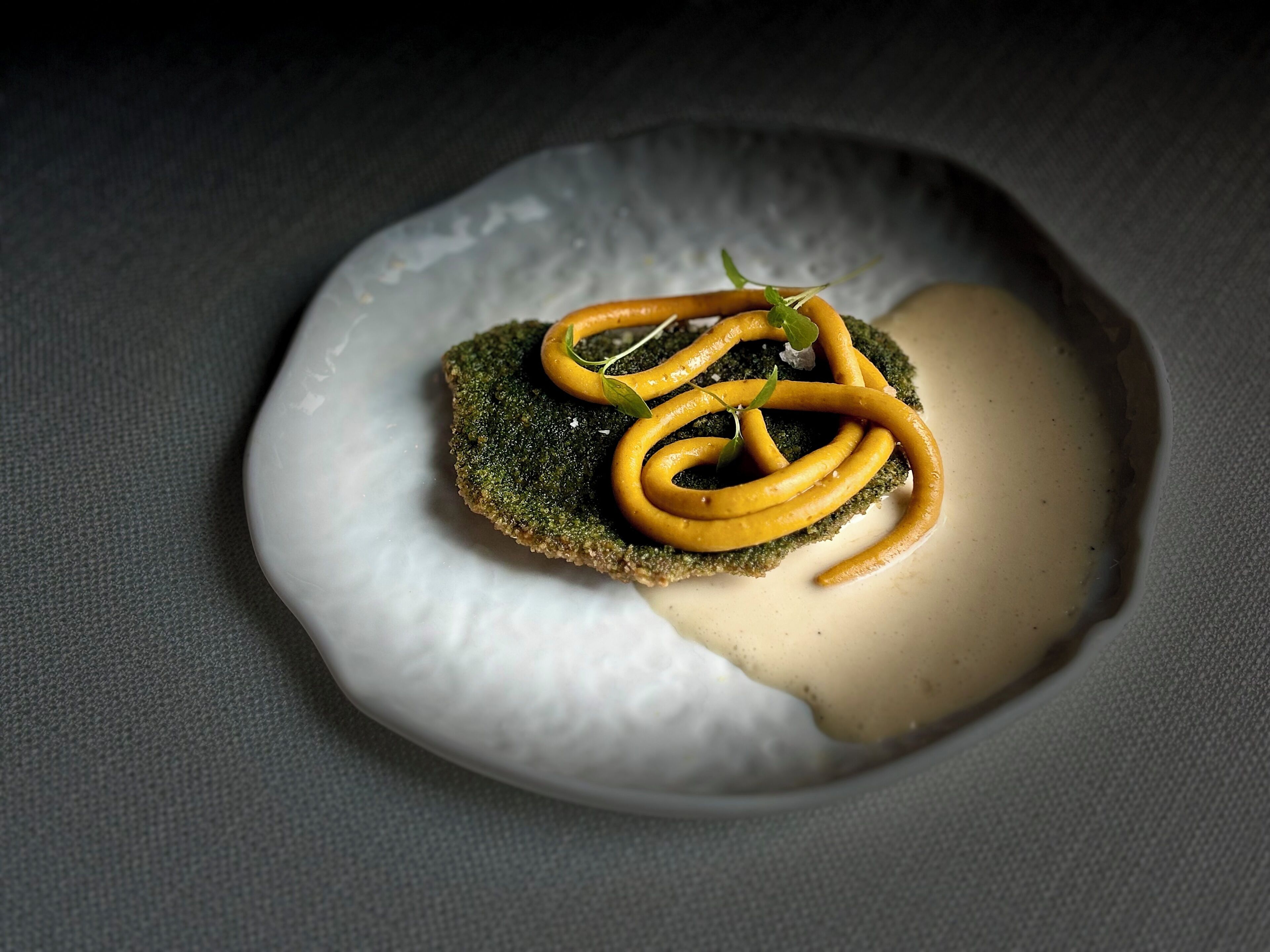 A plate with a green vegetable, possibly broccoli, topped with a creamy sauce and garnished with a yellow vegetable, likely a type of squash or carrot.