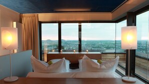 A modern hotel room with a large bed, two bedside lamps, and a view of a cityscape through large windows. - The Seven Hotel (Esch-sur-Alzette)
