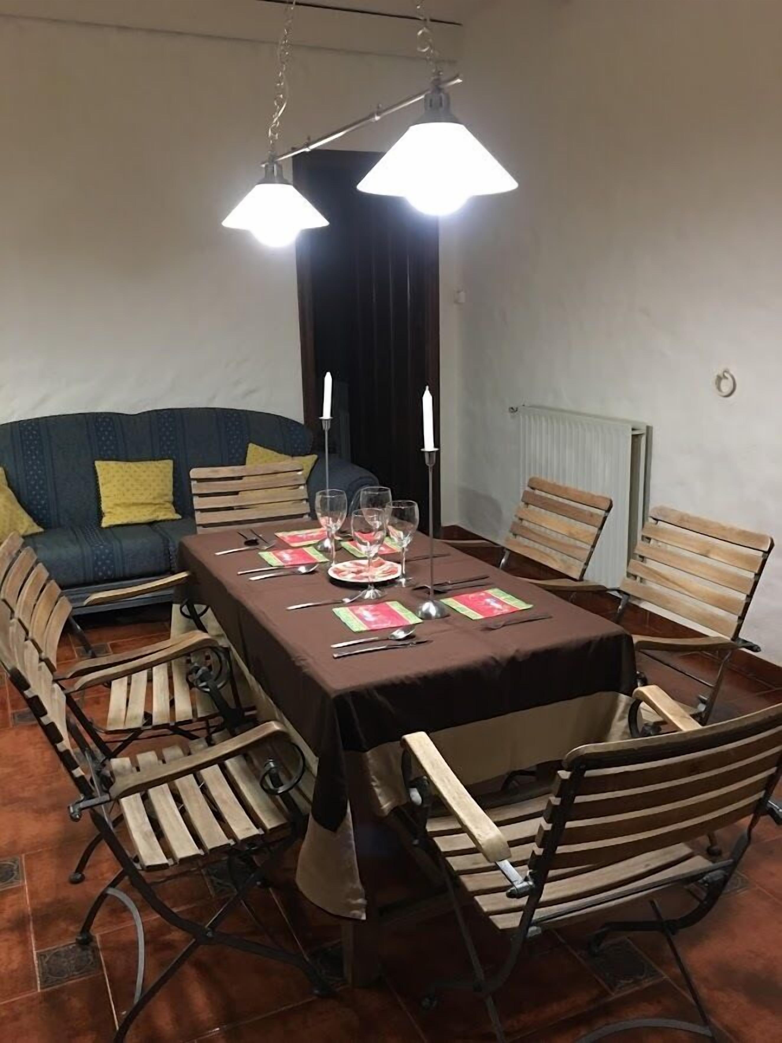 Comedor con una mesa lista para cuatro personas, rodeada de sillas de madera. Dos lámparas colgantes están sobre la mesa, y se ve un radiador en la pared a la derecha.