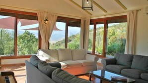 Amplia sala de estar con un sofá beige, un sofá gris y una mesa de centro. Amplias ventanas ofrecen vistas a zonas verdes y montañas.