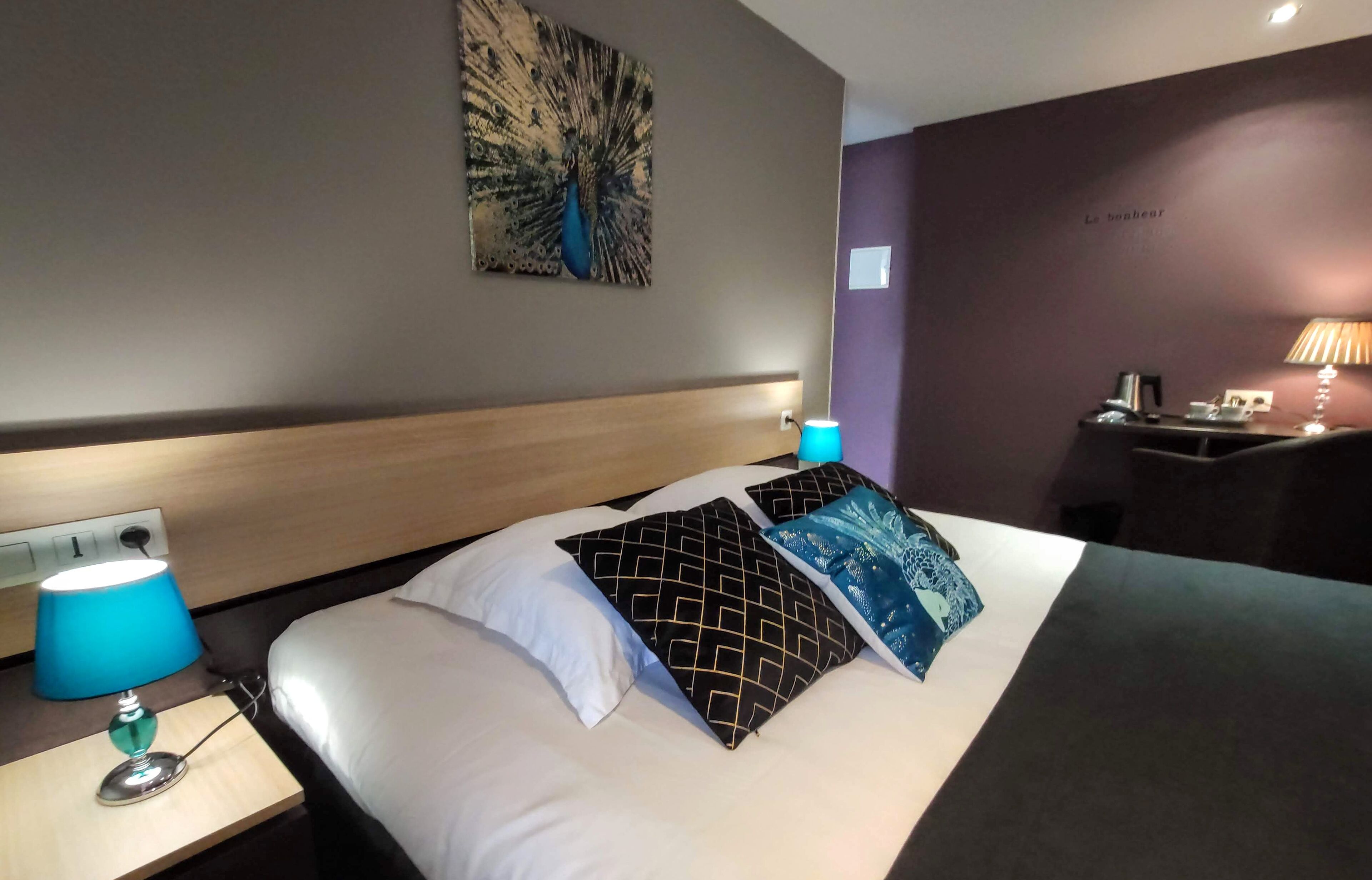 Habitación de hotel con cama, mesita de noche con lámpara, escritorio con lámpara y un cuadro de pavo real en la pared.