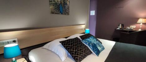 Habitación de hotel con cama, mesita de noche con lámpara, escritorio con lámpara y un cuadro de pavo real en la pared.