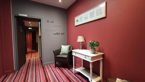 Ein Hotel-Flur mit rotem Teppich, einer grauen Wand mit einem Schild, einem schwarzen Sessel mit Kissen, einem weißen Konsolentisch mit Lampe und Pflanze sowie einem gerahmten Bild an der Wand.