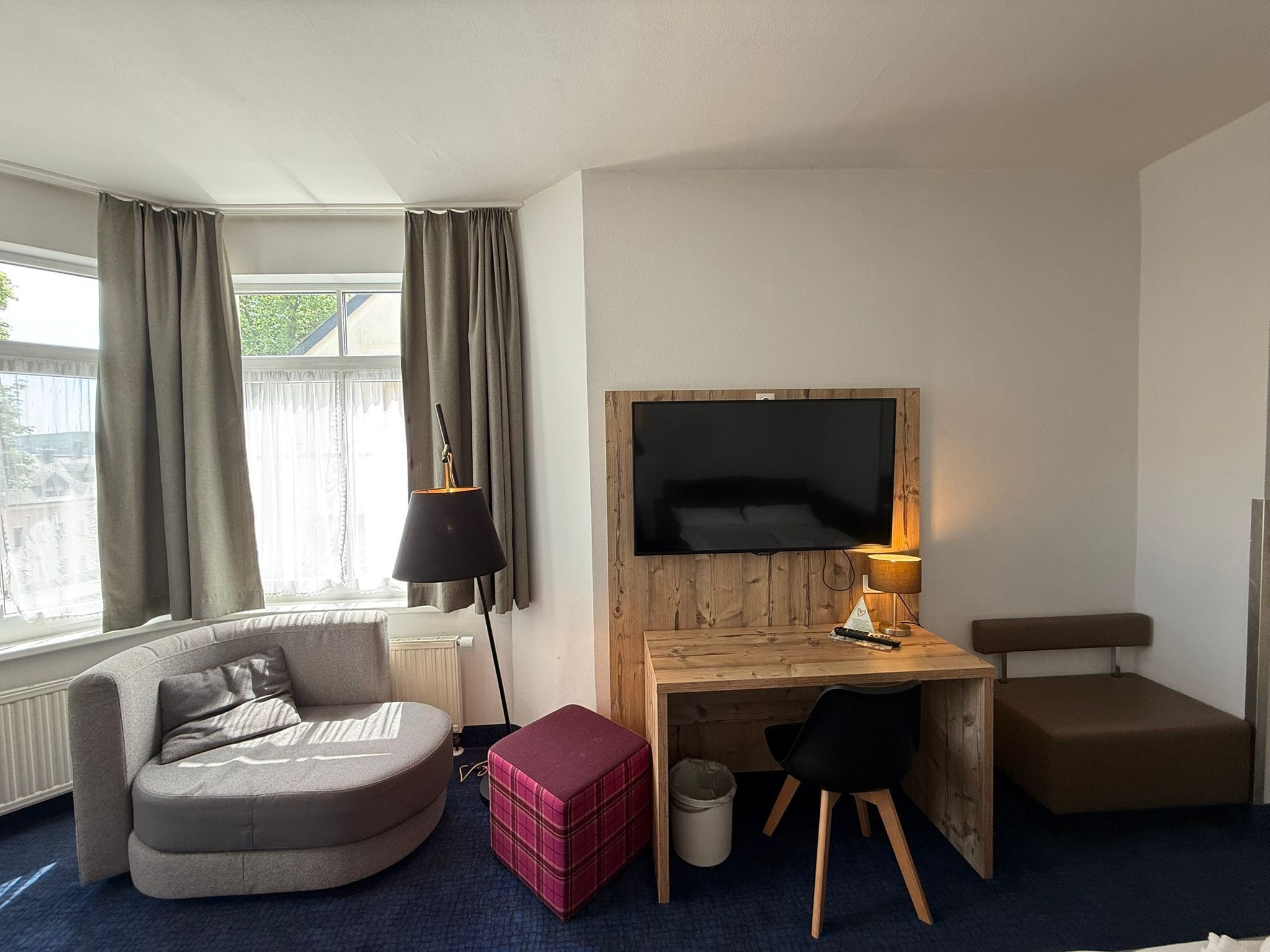 A modern hotel room with a sofa, a wooden desk with a TV, a lamp, and a small table.