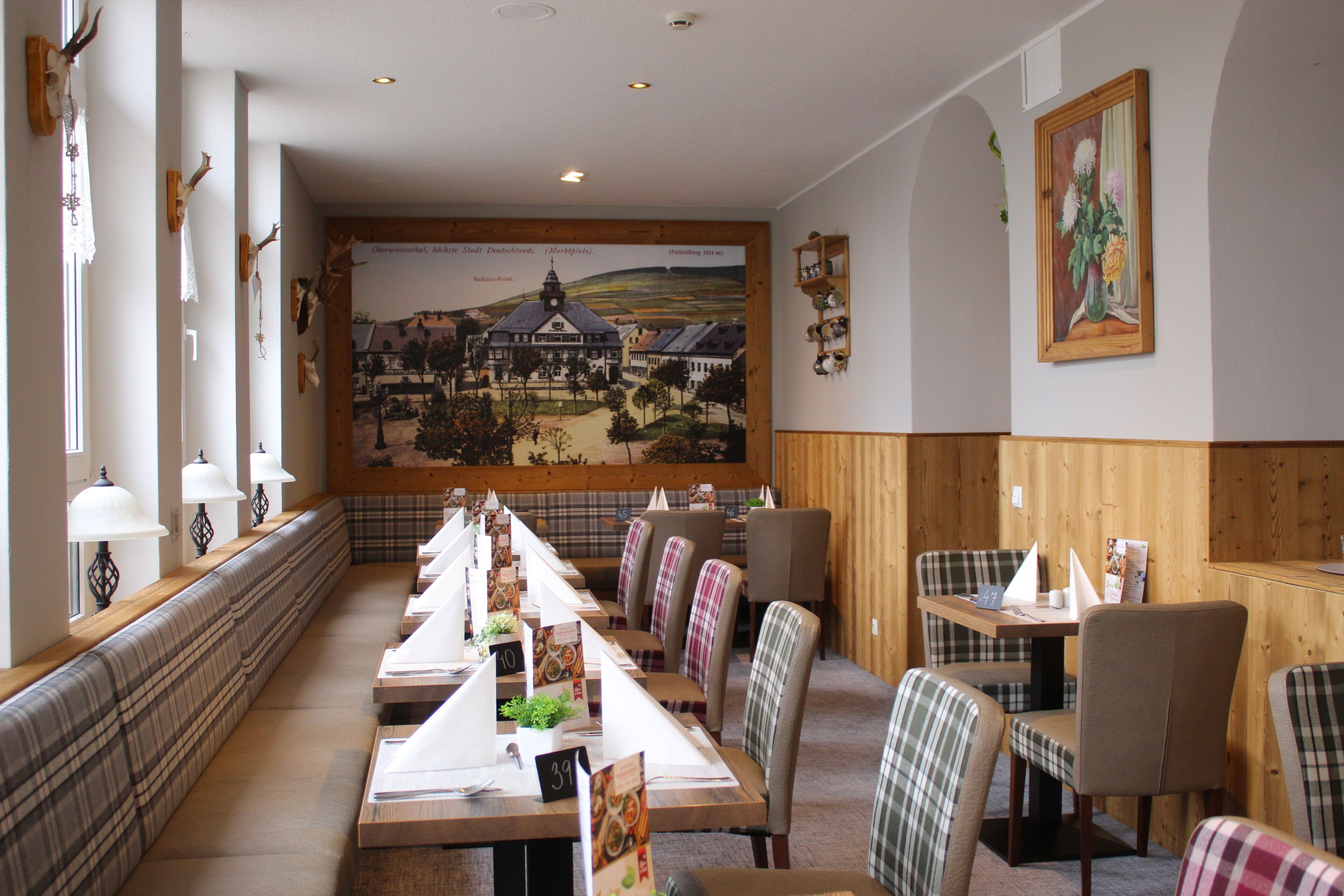 A restaurant with long communal tables, plaid chairs, and a mural of a town on the wall.
