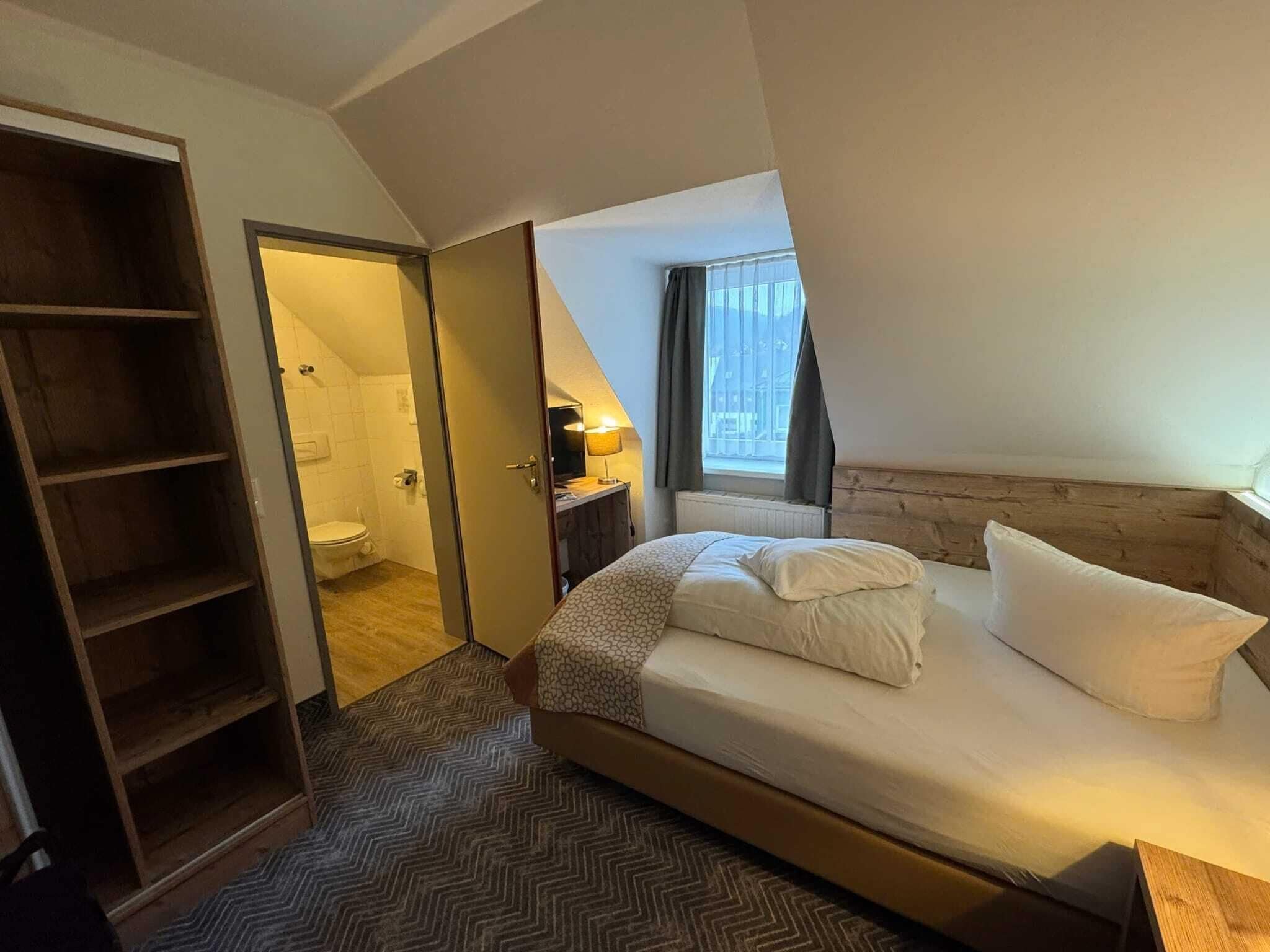 Une chambre à coucher avec un lit, une armoire en bois, une salle de bain avec des toilettes et une fenêtre avec des rideaux.