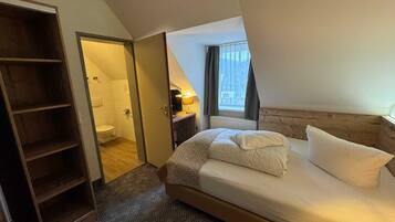 Une chambre à coucher avec un lit, une armoire en bois, une salle de bain avec des toilettes et une fenêtre avec des rideaux.