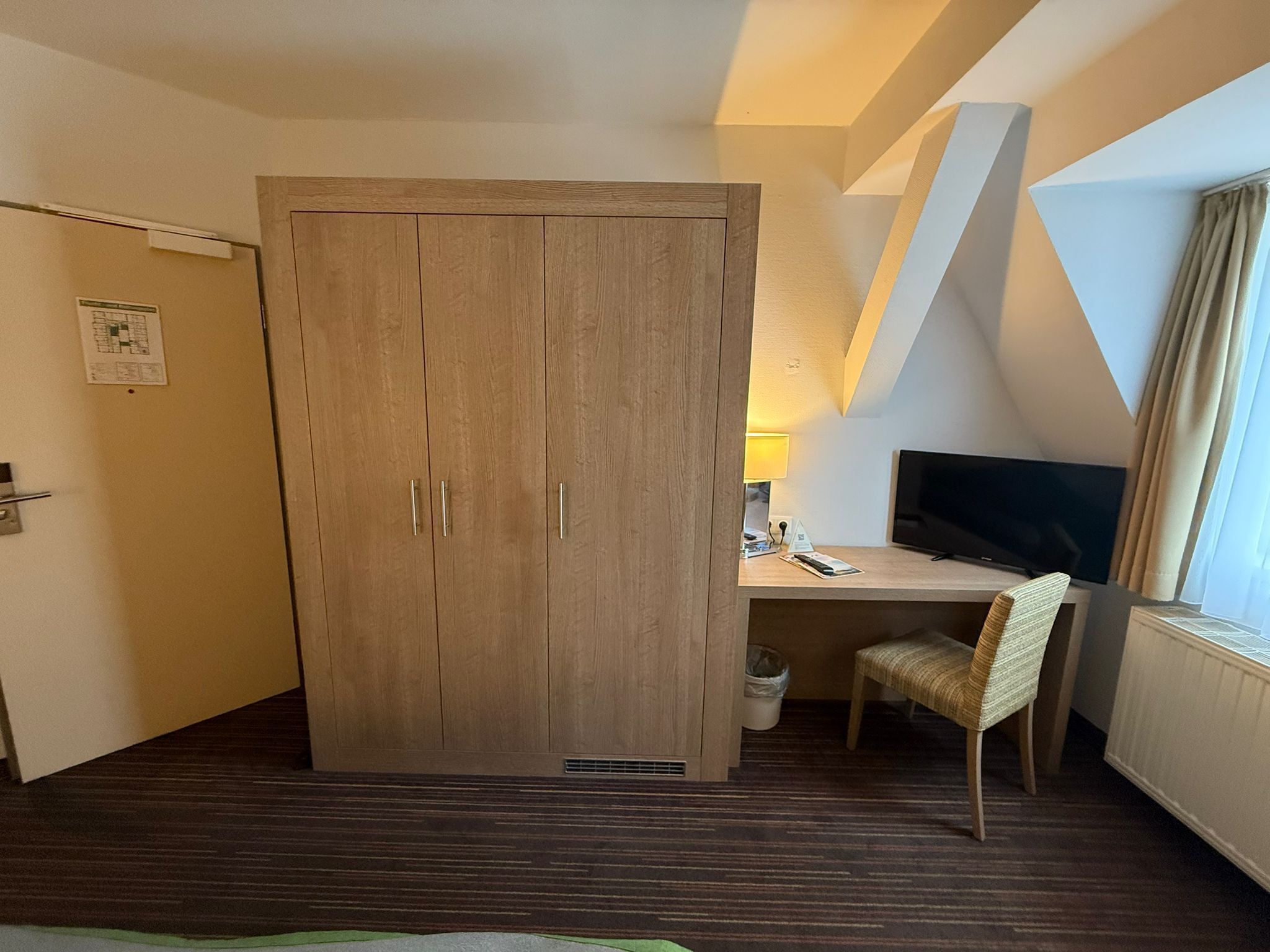 Een kamer met een houten garderobe, een bureau met stoel, een televisie en een raam met gordijnen.