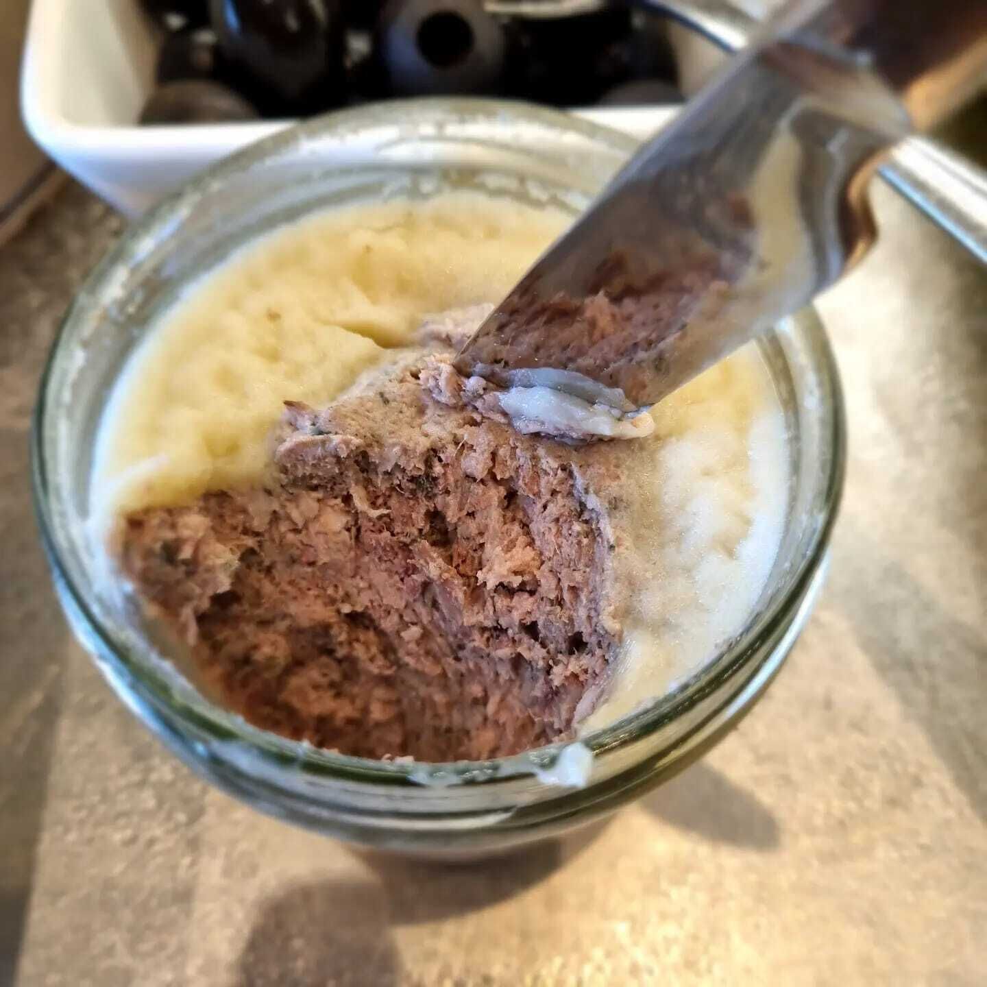 A glass bowl containing a mixture of mashed potatoes and a dark brown substance, possibly a spread or topping, with a knife in it.