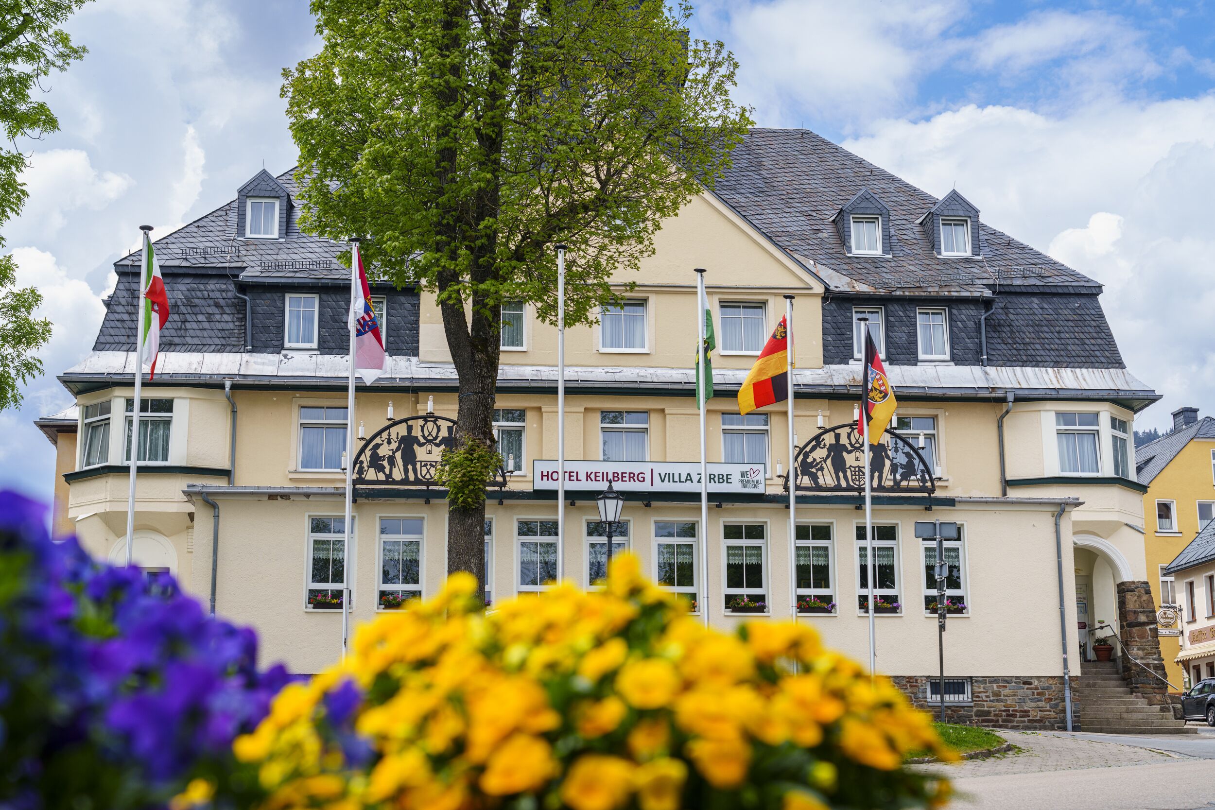 A hotel with multiple flags, a sign reading "HOTEL KEILBERG | VILLA ZRBE," and a tree in the foreground.