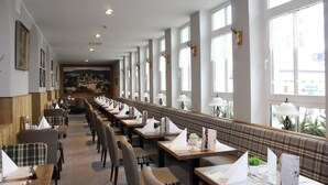 Un amplio comedor con ventanas altas, mesas dispuestas con manteles blancos y un mural en la pared.