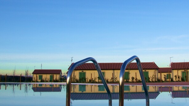 A swimming pool with metal ladders in the foreground, reflecting nearby buildings with red roofs and green shutters. - Eden Park Resort (San Giuliano Terme)