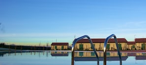 A swimming pool with metal ladders in the foreground, reflecting nearby buildings with red roofs and green shutters.