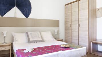 Ein ordentlich bezogenes Bett mit rosa Blumenmuster, zwei Nachttische mit Lampen, ein großer Kleiderschrank und ein Wandventilator.