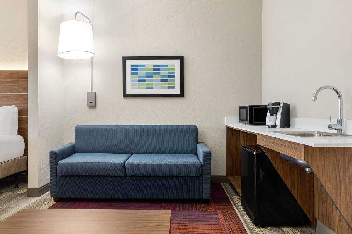 A modern hotel room with a sofa, a wall-mounted lamp, a framed abstract art piece, a kitchen area with a sink and appliances, and a patterned rug.