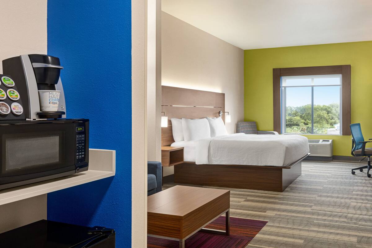 A hotel room with a bed, a desk, a microwave, a coffee maker, and a window with a view of trees.