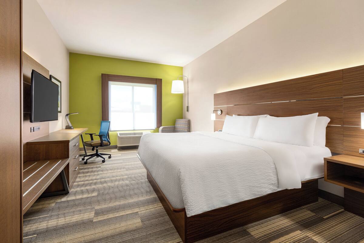 A hotel room with a large bed, a desk, a chair, a TV, and a window with natural light.