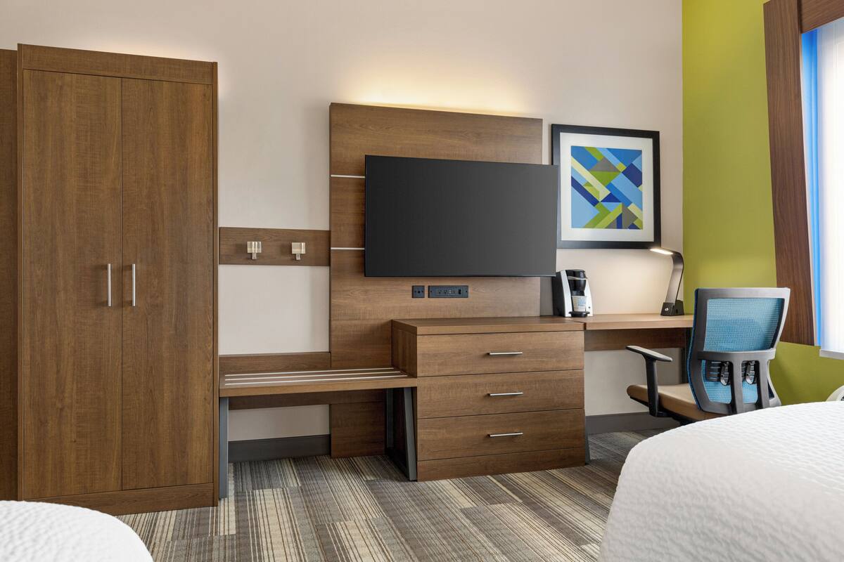 A hotel room with a wooden wardrobe, a desk with drawers, a flat-screen TV, a framed artwork, and a chair.