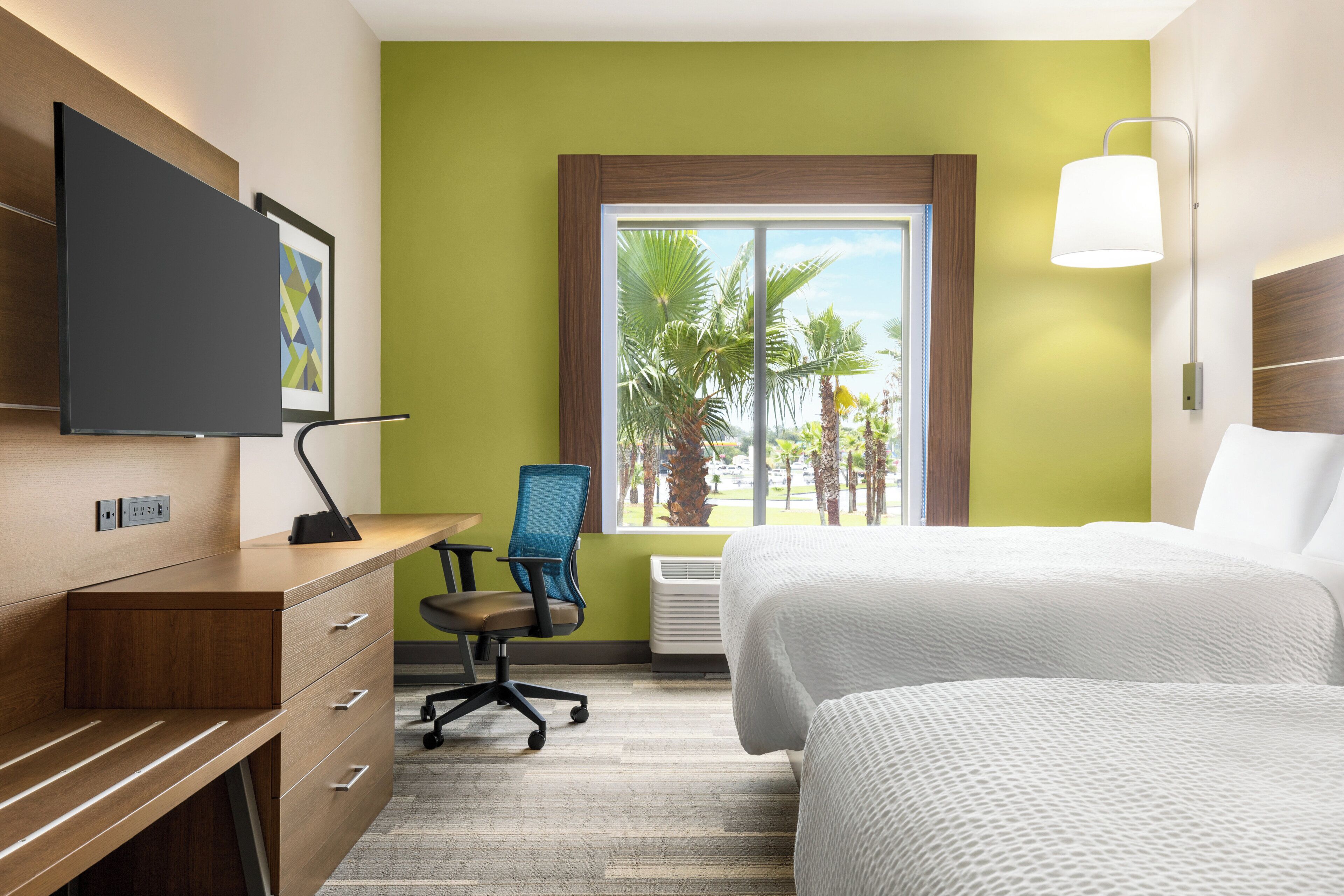 Una camera d'albergo con un letto, una scrivania con una sedia, una televisione e una finestra vista palme.