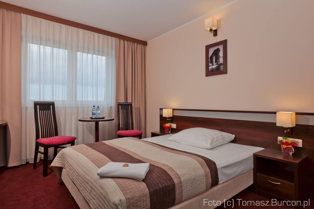 A hotel room with a large bed, two bedside tables, a chair, a small table, and a window with curtains.