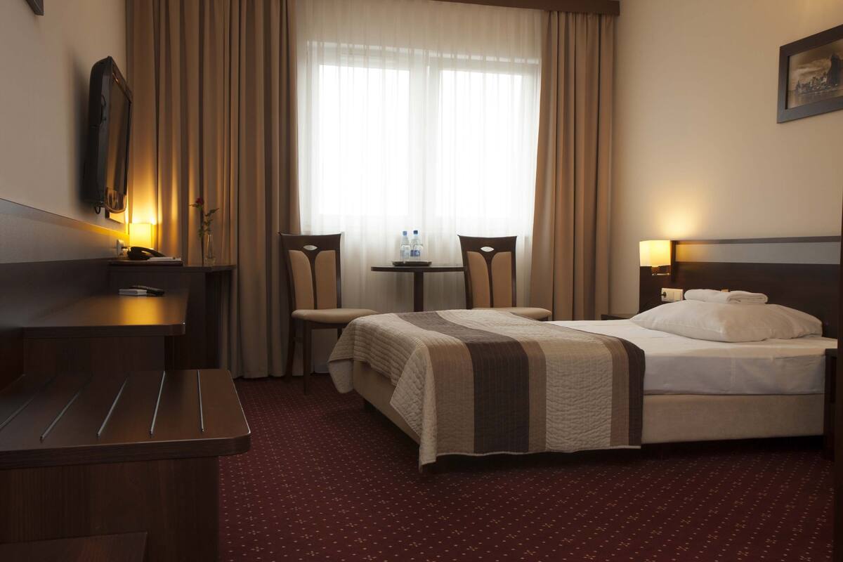 A hotel room with a bed, a desk, a chair, a small table with two bottles, and a TV on the wall.