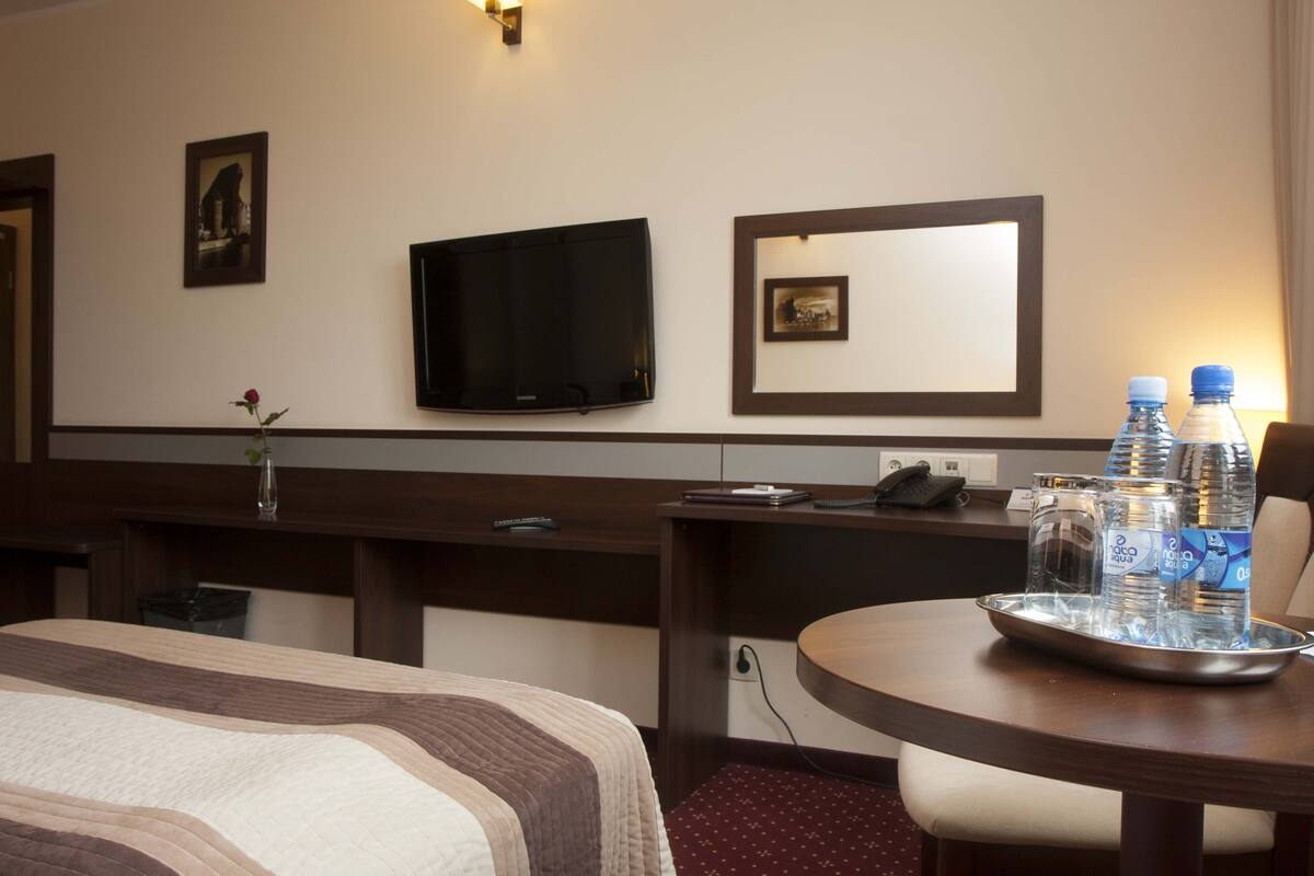 A hotel room with a bed, a desk, a television, a mirror, and a tray with water bottles.