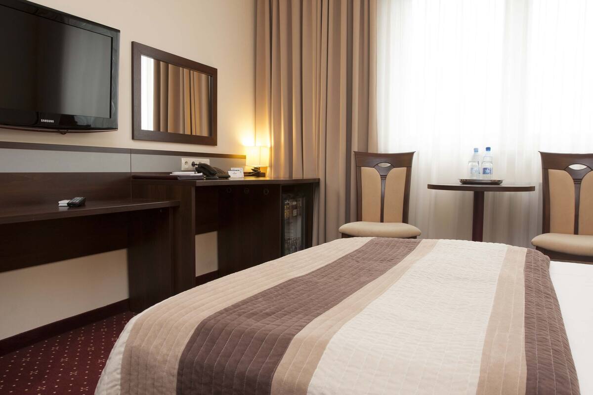 A hotel room with a bed, a desk, a chair, a TV, and a small table with water bottles.