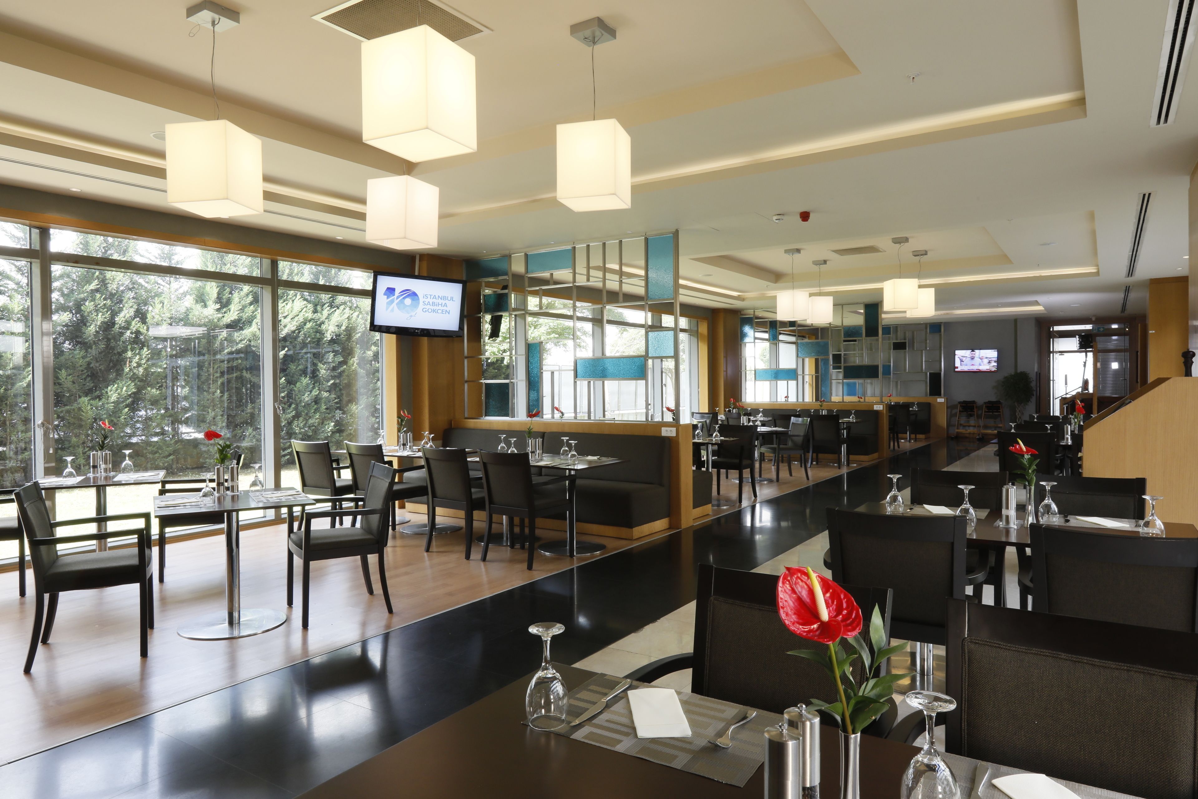 A modern restaurant with high ceilings, pendant lighting, and large windows offering a view of trees.