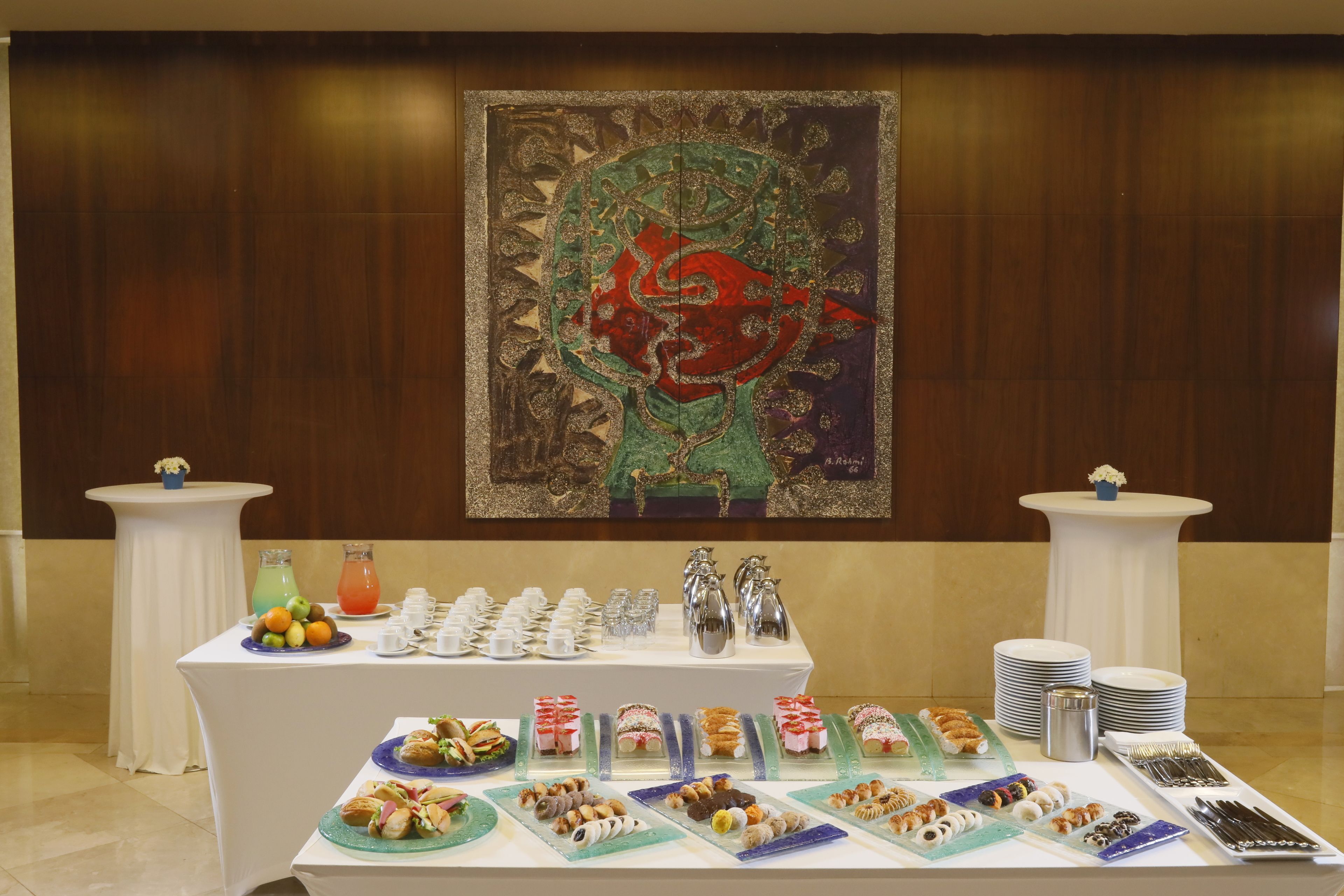 A buffet setup with various food items and beverages, a decorative wall painting, and two side tables with floral centerpieces.