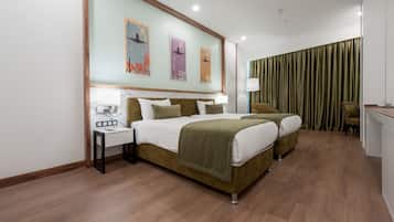 Quarto de hotel com uma cama grande, mesas de cabeceira, escrivaninha e uma cadeira. Há três quadros na parede e uma cortina na janela.