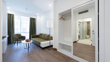 A modern hotel room with a wooden floor, a green sofa, a small round table with a vase of flowers, a lamp, and a walk-in closet with a mirror.