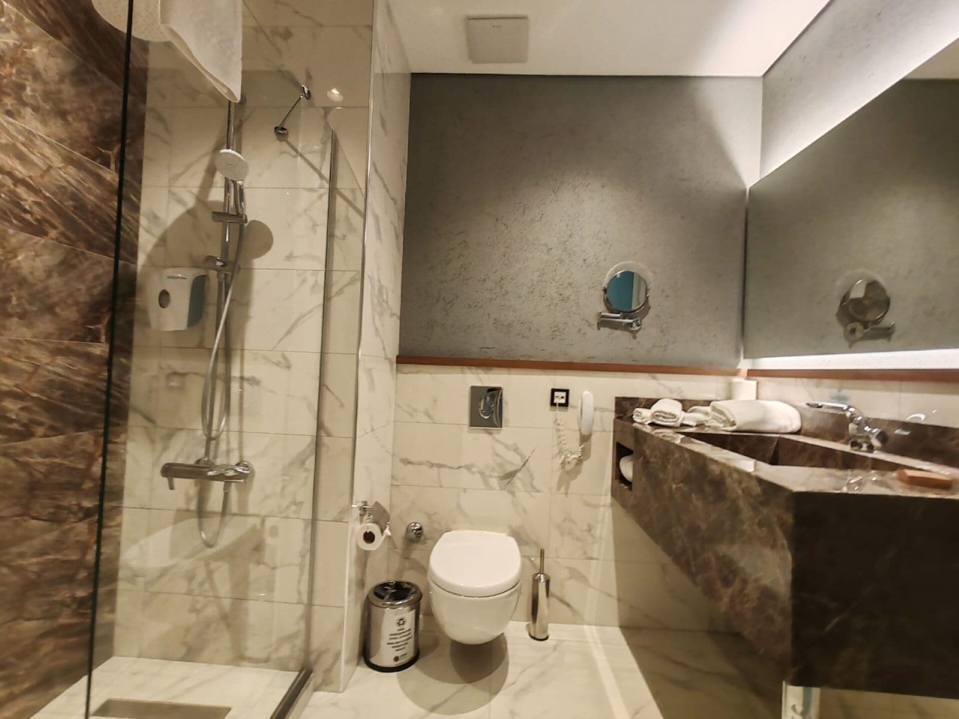 Ein modernes Badezimmer mit einer Duschkabine, einer Wand aus Marmorplatten, einer Toilette und einem Waschbecken mit Handtuchhalter.