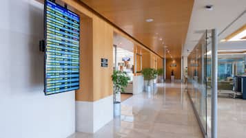 Un corridoio moderno di un hotel, con un pannello informativo digitale sui voli, piante in vaso e una parete divisoria in vetro.