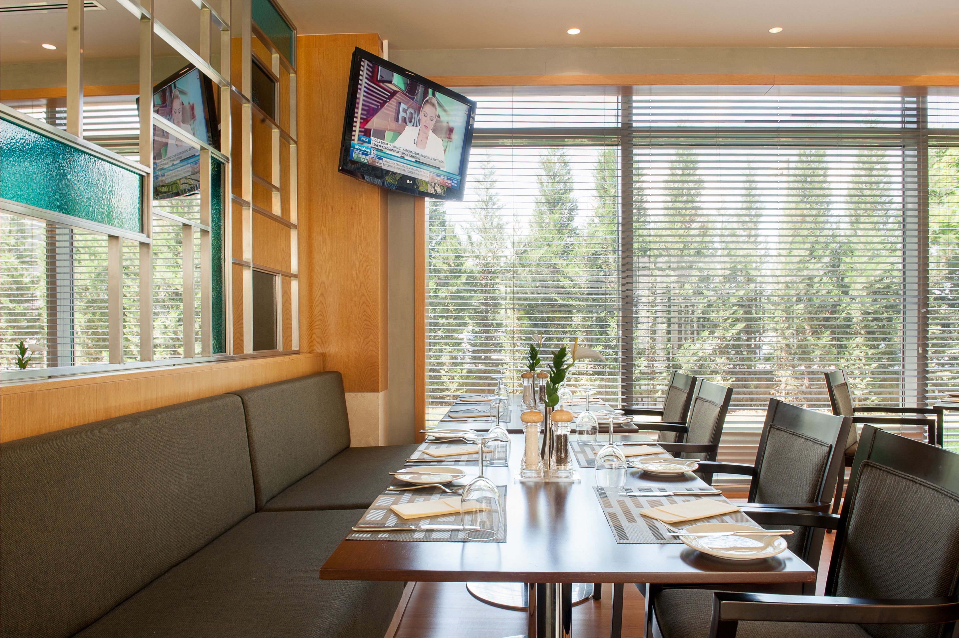 A dining area with a long table set for a meal, a television mounted on the wall, and large windows with blinds.