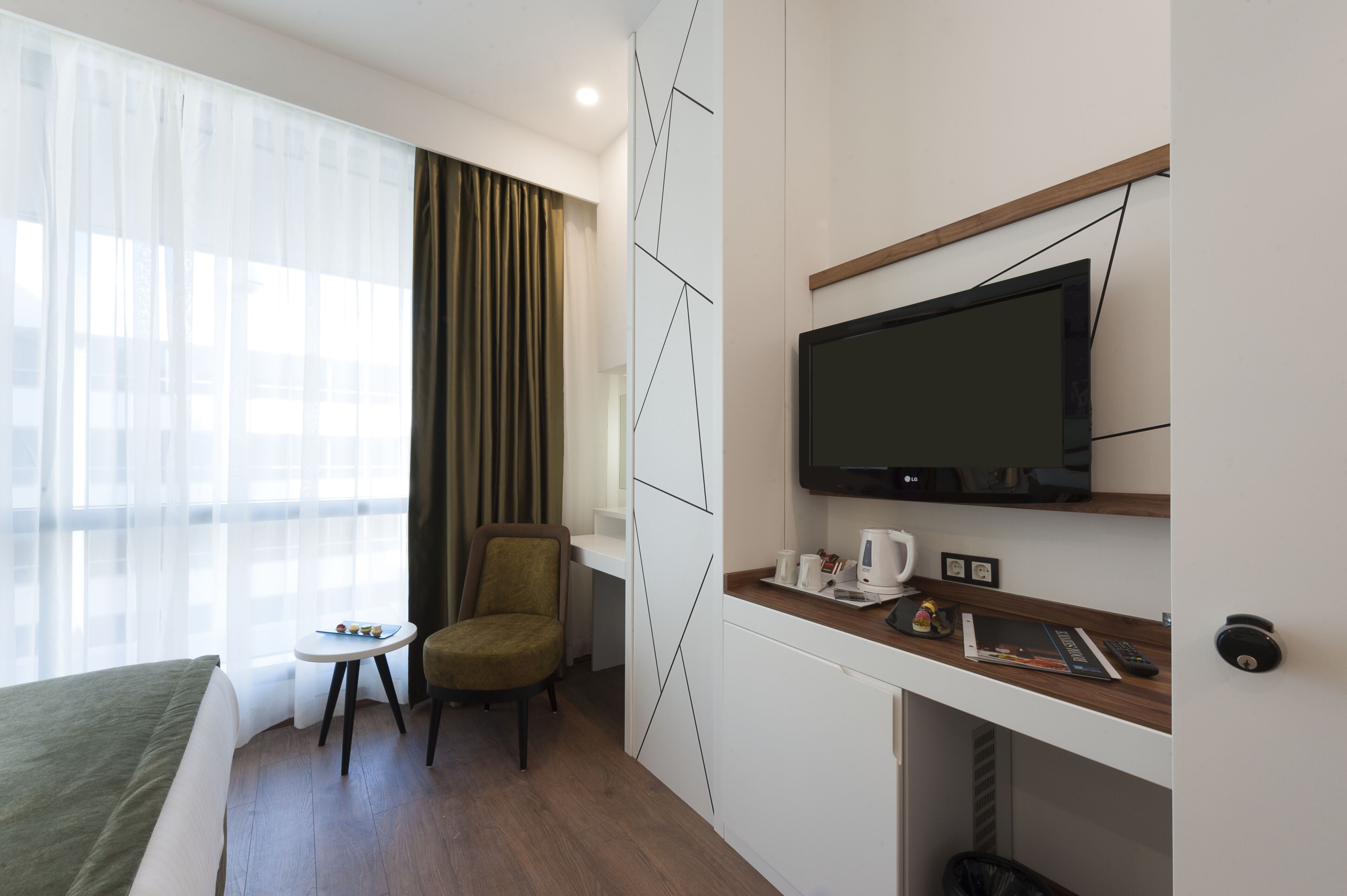 Habitación de hotel con una cama, una mesita con una silla, un televisor en la pared y una ventana con cortinas.