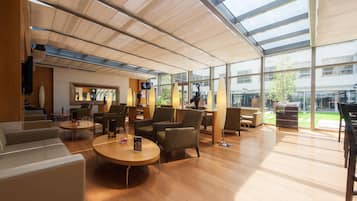Een ruime lobby met een hoge plafond, grote ramen en diverse zithoekjes, waaronder banken en fauteuils.